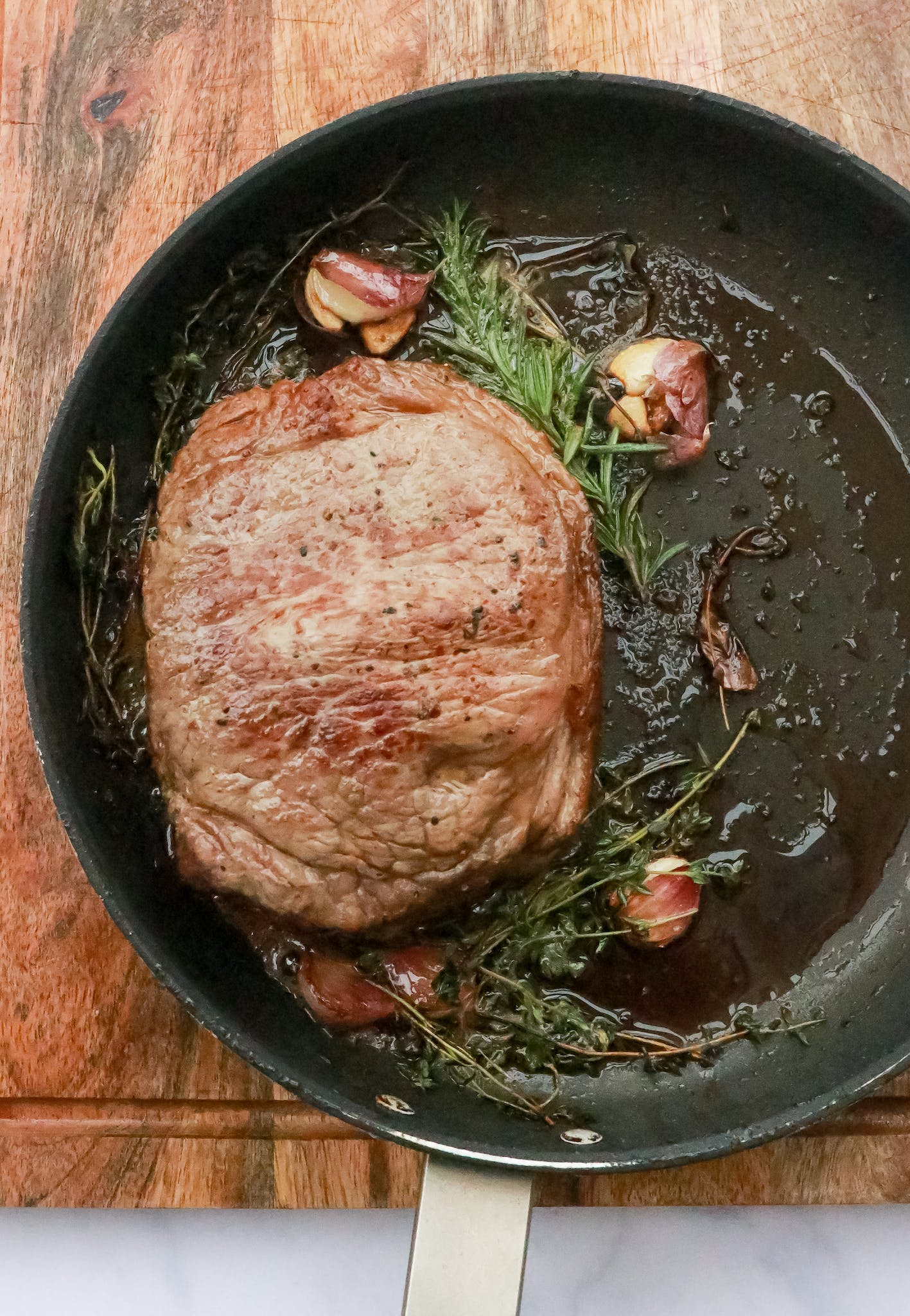 Top view of piece of meat steak in hot pan with rosemary and garlic placed on wooden cutting board in kitchen